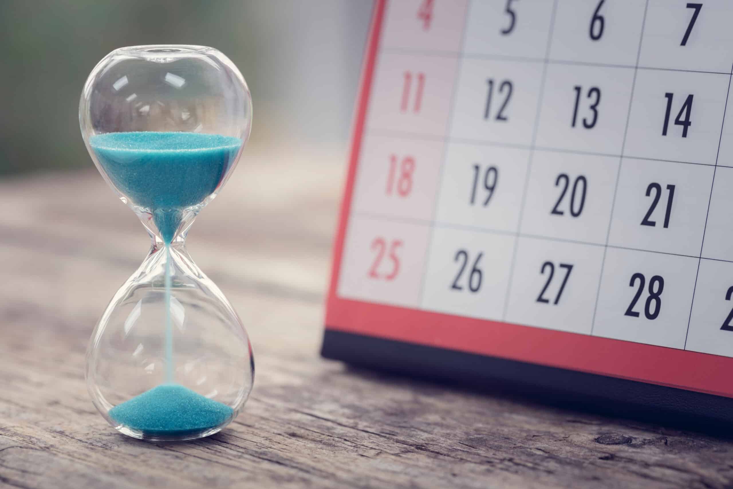 A sand timer is on a desk with a calendar.