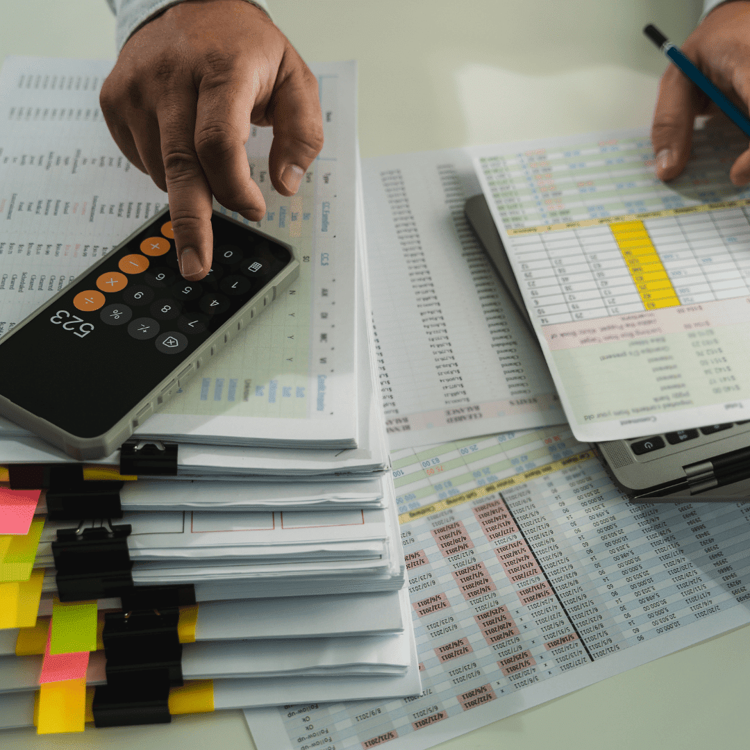 An accountant is seated with a pen, calculator app on his phone and financial documents working out figures.