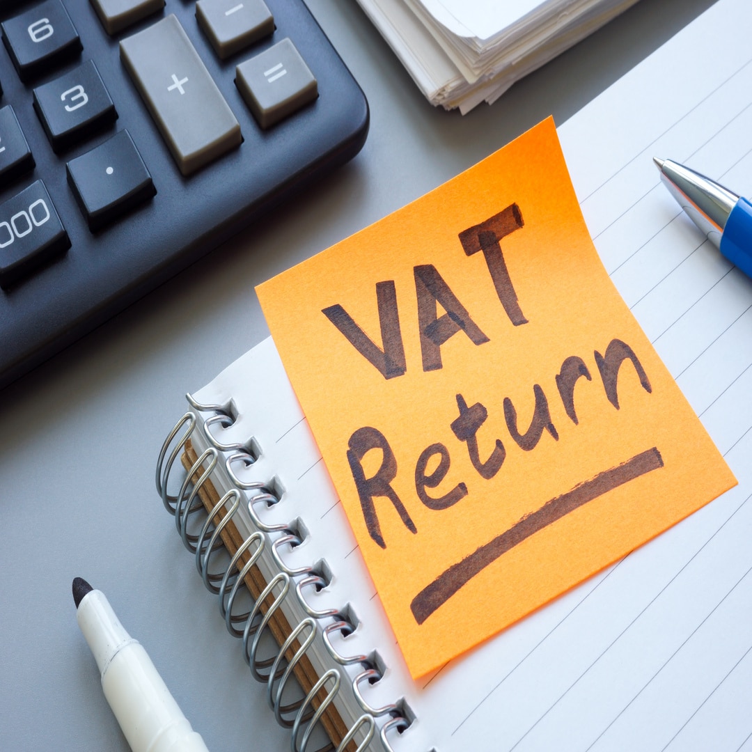 Image of a notebook, pens and paperwork, with an orange sticky note on the open notebook, with 'VAT Return' written onto the sticky note.
