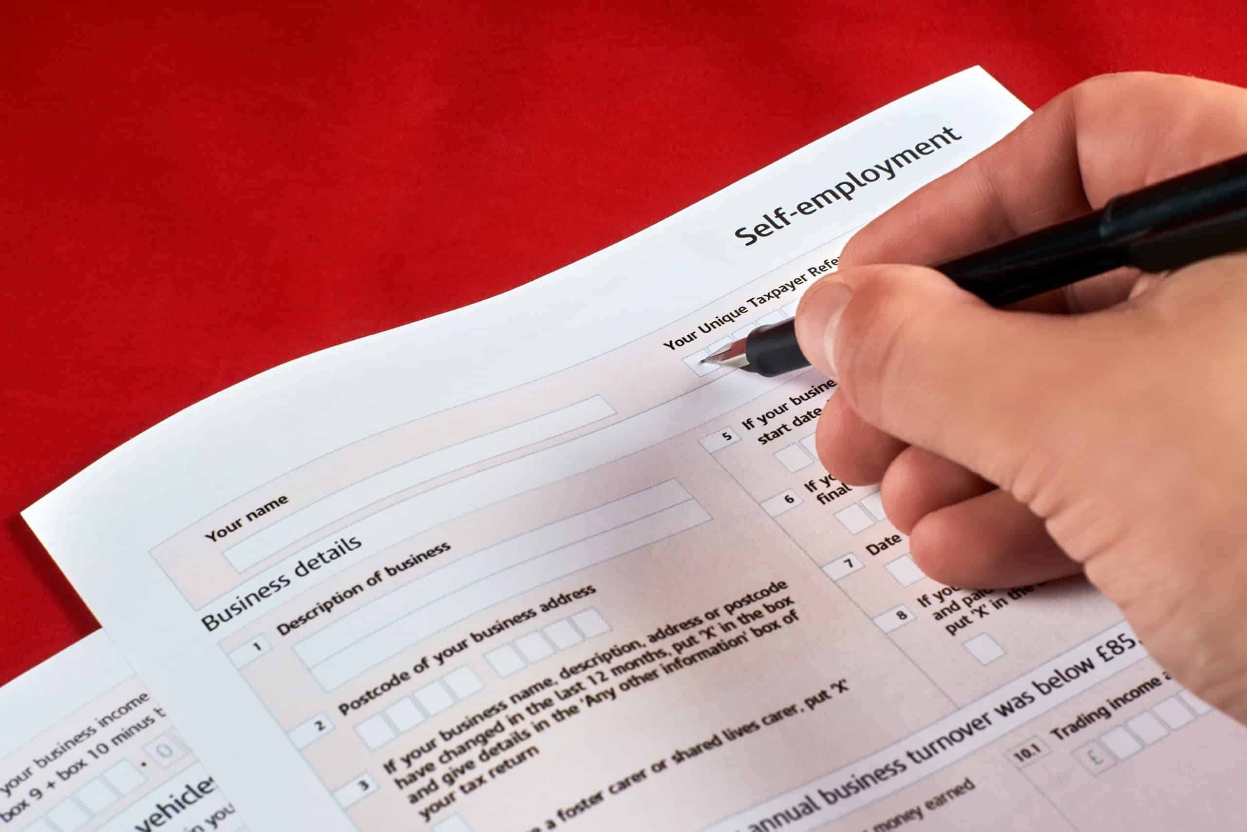 A person preparing to fill out a self-employment assessment form on a desk with a pen in hand.